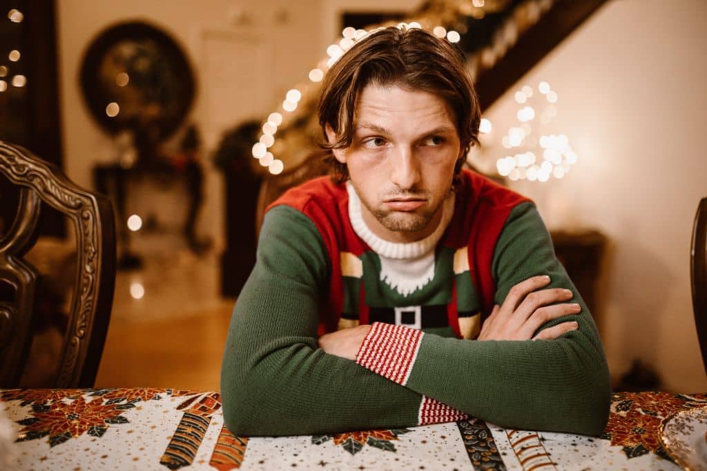 A man sat at a Christmas dinner table wearing a festive jumper looking sullen - Which Rehab