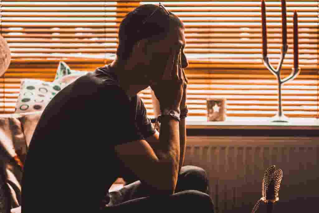 A man holding his head in his hands in a darkened room contemplating rehab - Which Rehab